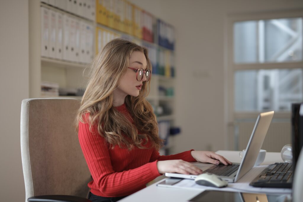 Mujer rubia con lentes rojos que traba remotamente en un laptop dentro de una habitación